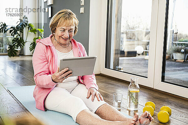 Lächelnde blonde Seniorin mit Tablet-PC sitzt zu Hause auf einer Trainingsmatte