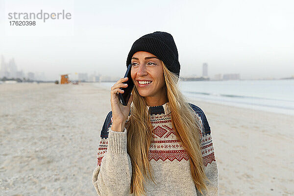 Lächelnde Frau mit schwarzer Strickmütze telefoniert am Strand