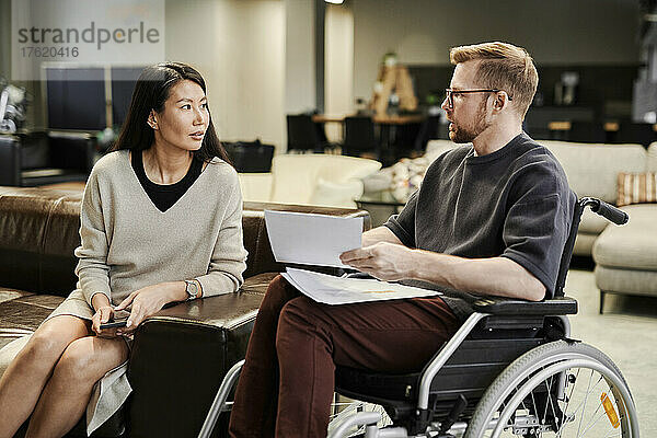 Behinderter Kollege mit Dokumenten im Gespräch mit Geschäftsfrau  die auf dem Sofa im Büro sitzt