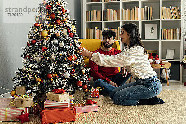 Junger Mann sitzt neben seiner Freundin und schmückt zu Hause den Weihnachtsbaum