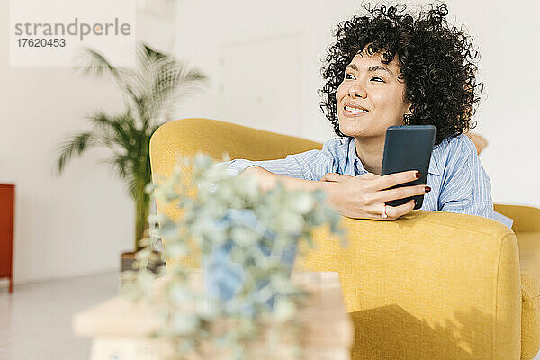 Smiling young woman lying on sofa with smart phone at home