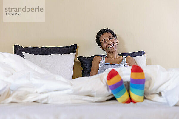 Lächelnde Frau mit bunten Socken sitzt zu Hause im Bett