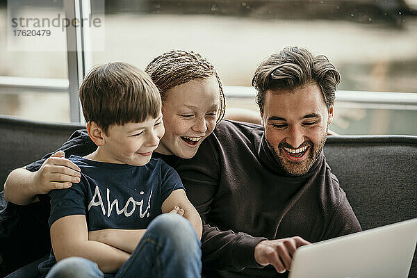 Glücklicher Mann zeigt auf Laptop und sitzt mit Söhnen zu Hause