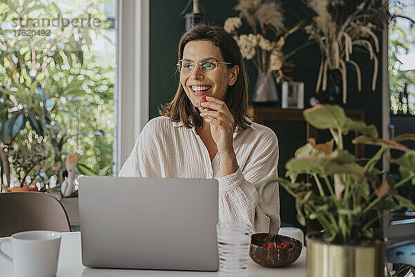 Lächelnder Freiberufler mit Laptop  der Erdbeeren isst und im Heimbüro arbeitet