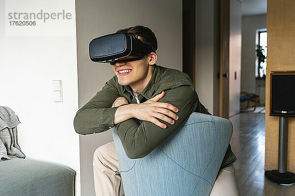Glücklicher junger Mann mit Virtual-Reality-Simulator  der sich zu Hause auf einen Stuhl lehnt