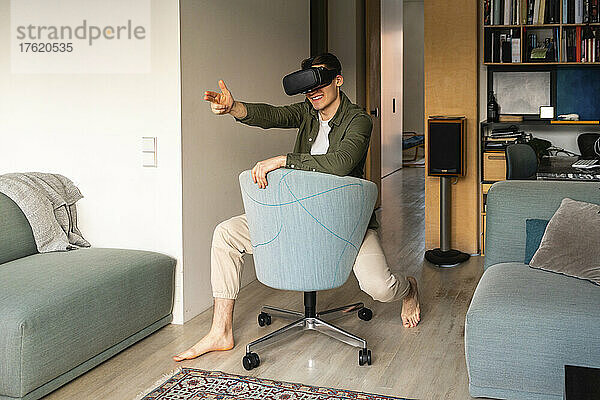Lächelnder junger Mann mit Virtual-Reality-Simulator gestikuliert auf einem Stuhl zu Hause