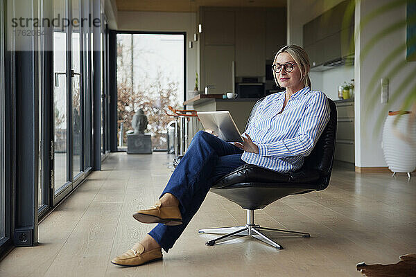 Frau mit Brille und Tablet-PC sitzt zu Hause auf einem Stuhl