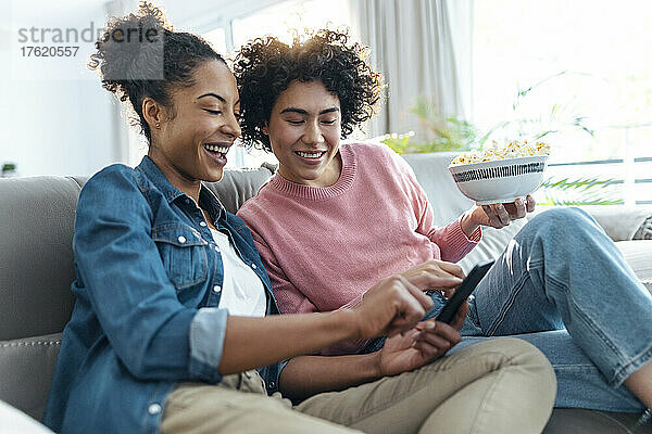 Glückliche Frau  die ihr Smartphone mit einer Freundin teilt  die auf dem Sofa im Wohnzimmer sitzt