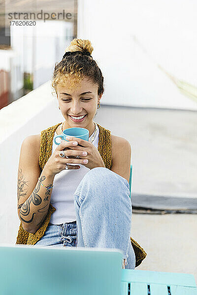 Happy young woman holding tea cup sitting with laptop at table on rooftop