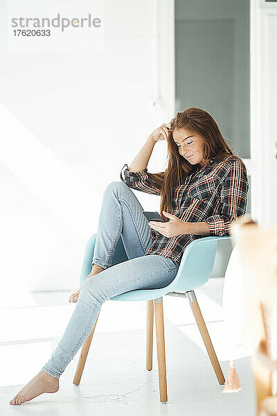 Junge Frau sitzt zu Hause auf einem Stuhl und benutzt ein Smartphone