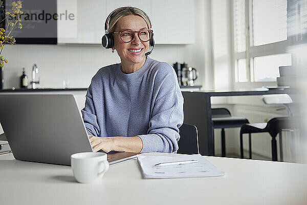 Glückliche blonde Geschäftsfrau mit Headset sitzt mit Laptop am Tisch