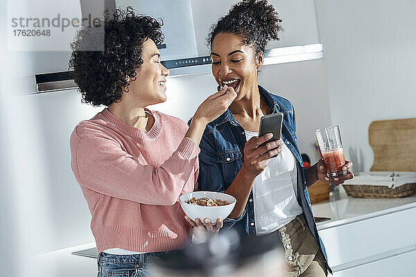 Junge Frau füttert Freund  der zu Hause in der Küche steht  mit Müsli