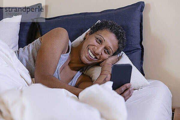 Lächelnde Frau  die im Schlafzimmer auf dem Bett liegt und ihr Mobiltelefon benutzt