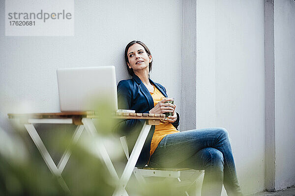 Geschäftsfrau mit Laptop und Trinkglas sitzt auf Stuhl im Straßencafé