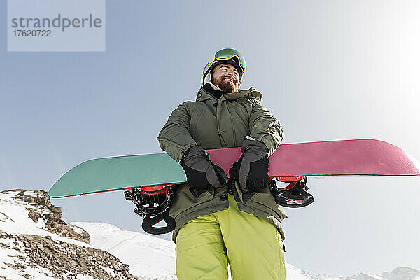 Lächelnder junger Mann steht an einem sonnigen Tag mit Snowboard