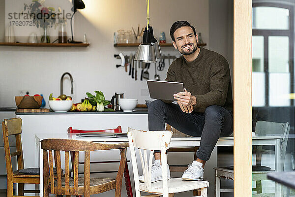 Lächelnder junger Mann mit Tablet-PC sitzt zu Hause am Tisch in der Küche