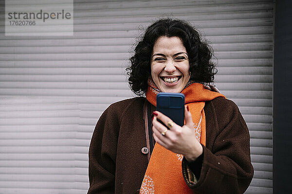 Glückliche junge Frau mit Mobiltelefon steht vor einem Wellpappenfensterladen