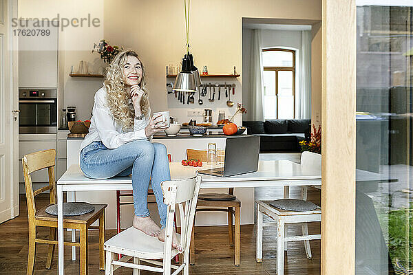 Glückliche junge Frau hält Kaffeetasse in der Küche und sitzt mit der Hand am Kinn