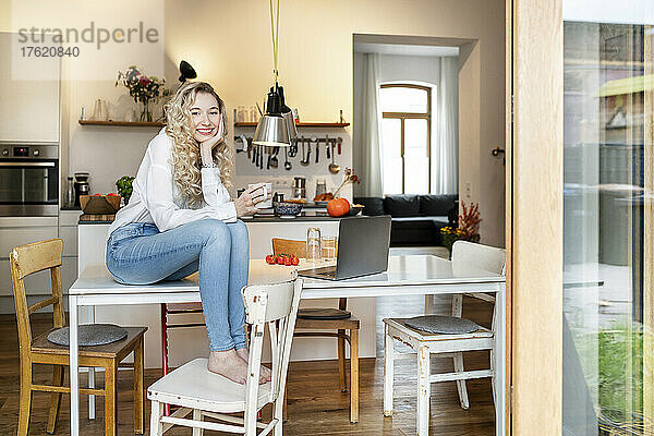 Glückliche blonde Frau hält eine Kaffeetasse und sitzt mit dem Kopf in der Hand auf dem Tisch