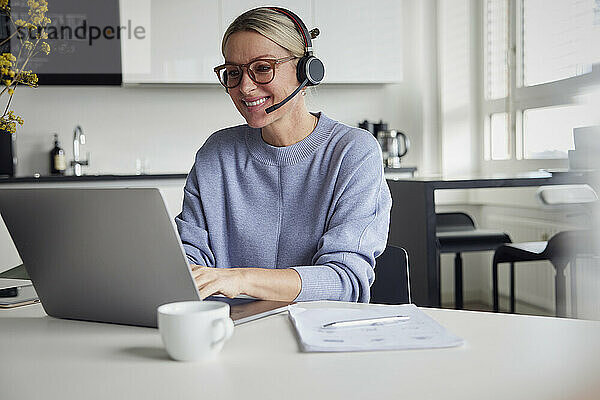Lächelnde Geschäftsfrau mit Headset und Laptop am Tisch sitzend