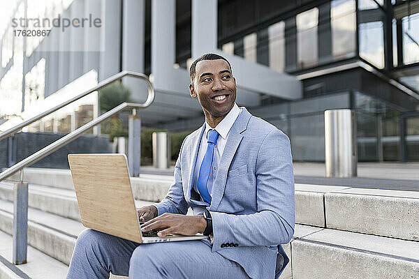 Lächelnder Geschäftsmann mit Laptop sitzt auf Stufen vor dem Gebäude