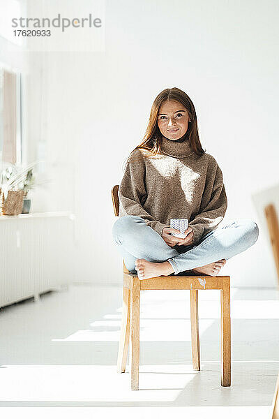 Junge Frau mit Kaffeetasse sitzt zu Hause auf einem Stuhl