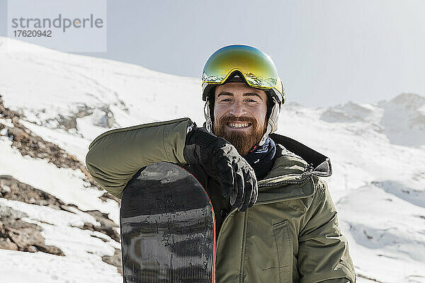 Lächelnder junger Mann mit Skibrille steht an einem sonnigen Tag mit Snowboard