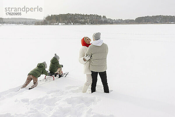 Paar umarmt sich  während Söhne im Winter im Schnee rodeln