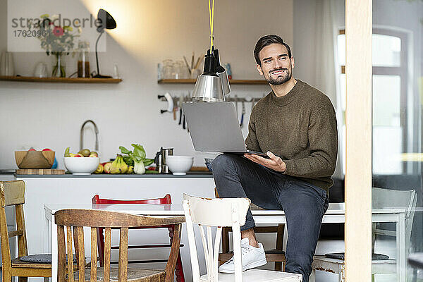 Lächelnder Freiberufler mit Laptop sitzt zu Hause auf dem Tisch in der Küche
