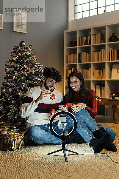 Glückliches Paar hält Weihnachtsschmuck in der Hand und vloggt zu Hause per Handy