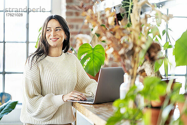 Lächelnde Geschäftsfrau steht mit Laptop zu Hause neben Topfpflanzen