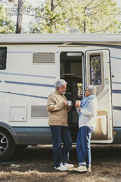 Älteres Paar mit Kaffeetasse steht neben Wohnmobil im Wald