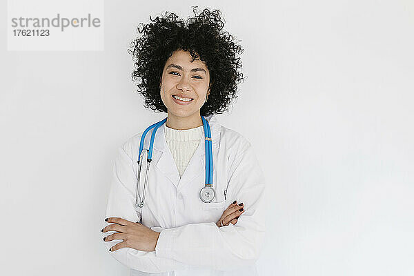 Lächelnder Arzt steht mit verschränkten Armen vor einer weißen Wand