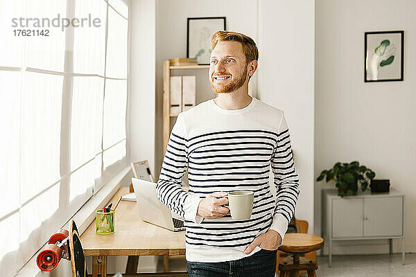Lächelnder junger Mann mit Kaffeetasse und der Hand in der Tasche zu Hause