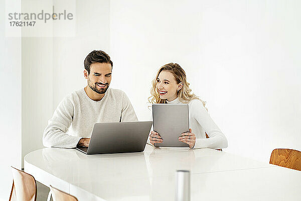 Lächelnde blonde Frau mit Tablet-PC blickt Kollege am Laptop an
