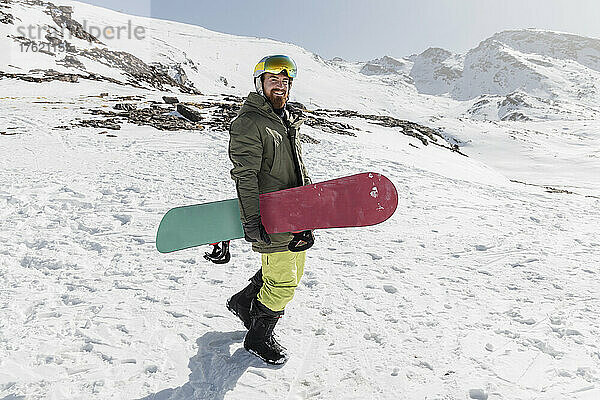 Lächelnder junger Mann  der an einem sonnigen Tag mit dem Snowboard spaziert
