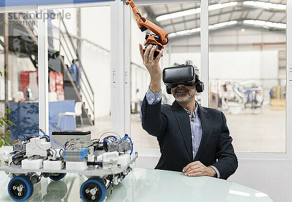 Lächelnder Geschäftsmann mit virtueller Realität und Roboterarmmodell am Schreibtisch