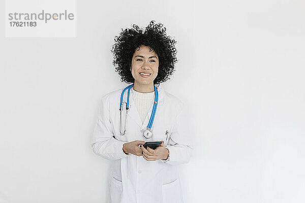 Lächelnder Arzt mit Mobiltelefon vor weißer Wand