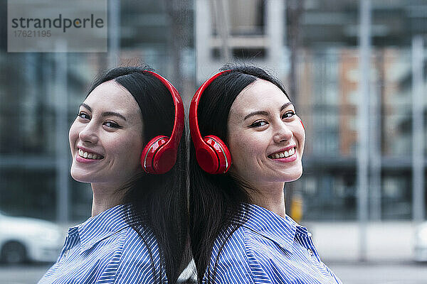 Smiling woman listening music through headphones leaning on glass wall