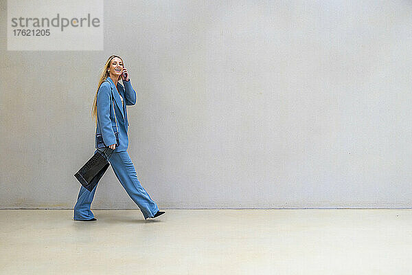Lächelnde Geschäftsfrau mit Einkaufstasche  die auf dem Smartphone spricht und an der Wand vorbeigeht