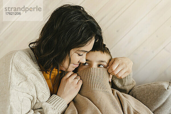 Lächelnde Mutter bedeckt Jungengesicht zu Hause mit Pullover