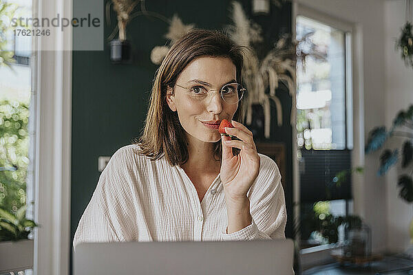 Berufstätige Frau mit Brille hält Erdbeere im Heimbüro
