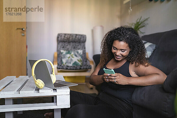 Lächelnde Frau  die SMS über ihr Smartphone sendet und zu Hause auf dem Sofa sitzt