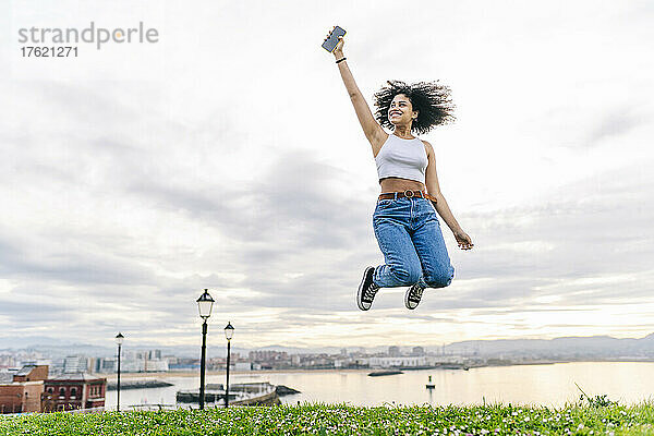 Fröhliche junge Frau mit Smartphone springt in der Natur