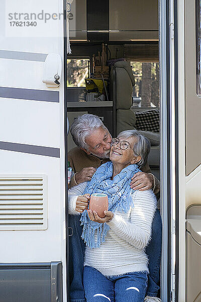Älterer Mann küsst glückliche Frau auf die Wange  die an der Tür des Wohnmobils sitzt