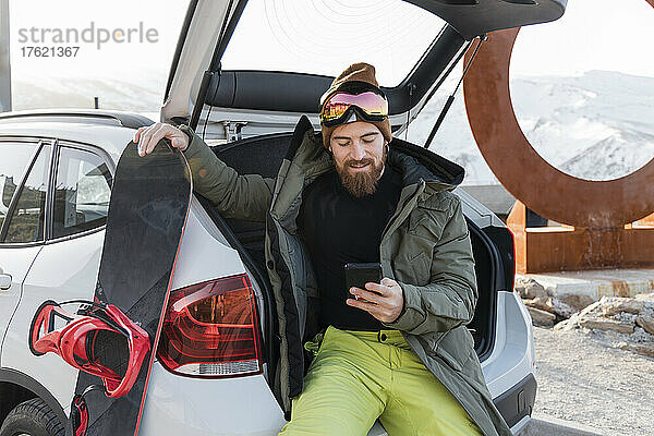 Junger Mann mit Snowboard sitzt im Kofferraum eines Autos und benutzt Smartphone