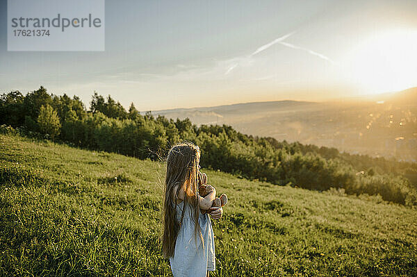 Kleines Mädchen mit Stofftier steht bei Sonnenuntergang auf der Wiese