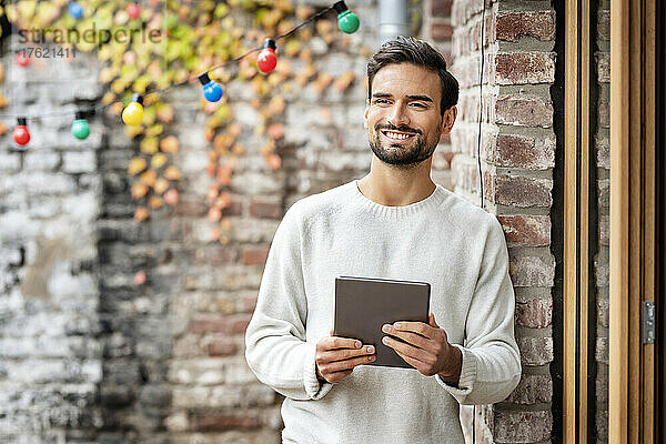 Lächelnder junger Mann mit Tablet-PC  der an einer Ziegelwand steht