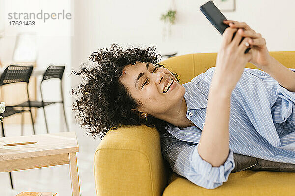 Glückliche junge Frau  die zu Hause auf dem Sofa liegt und Textnachrichten über ihr Smartphone sendet