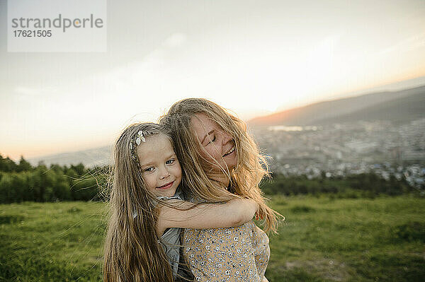 Süßes Mädchen mit blonden Haaren umarmt Mutter bei Sonnenuntergang